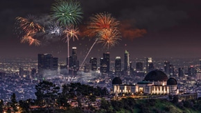 Colorful fireworks exploding over the Downtown Los Angeles skyline at night, with the Griffith Observatory illuminated in the foreground.